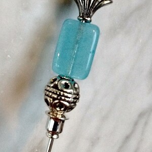 Può includere: Uno spillo d'argento con una perla di vetro blu e una parte superiore decorativa in argento. La parte superiore ha la forma di una conchiglia.