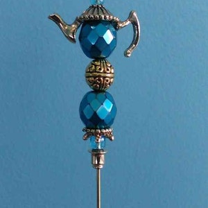 Op de afbeelding: Een zilveren theepotbedel met blauwe en gouden kralen, bevestigd aan een zilveren pin. De bedel is een decoratief accent voor een theepot of andere decoraties in huis.
