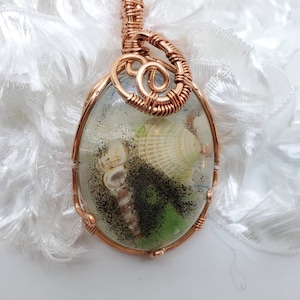 May include: A copper wire wrapped pendant with a clear resin oval containing seashells, sand, and green seaweed.
