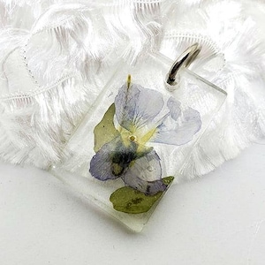 May include: A clear rectangular pendant with a pressed purple flower inside. The pendant has a silver loop at the top for attaching a chain.