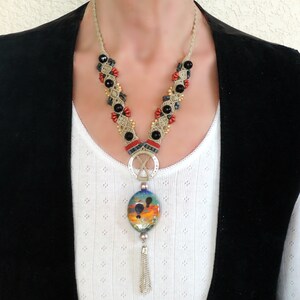 May include: A beige macrame necklace with black and red beads, a silver pendant with a colorful glass oval, and a silver tassel.
