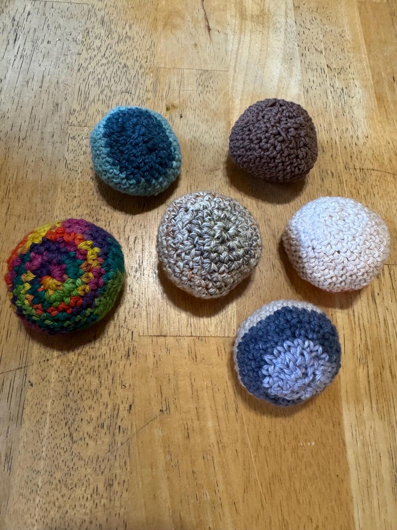 K&ouml;nnte beinhalten: Sechs handgefertigte H&auml;kelb&auml;lle in verschiedenen Farben sind auf einer Holzoberfl&auml;che angeordnet. Die B&auml;lle umfassen Farbt&ouml;ne von Blau, Braun, Regenbogen, Beige, Wei&szlig; und Blau-Grau. Die B&auml;lle sind rund und haben ein strukturiertes Aussehen.