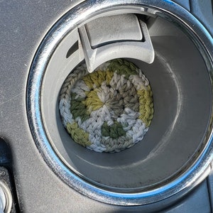 May include: A round, crocheted coaster in a car's cup holder. The coaster features a mix of green, white, and gray yarn, creating a textured, handmade appearance. The cup holder is silver and gray.