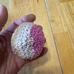 K&ouml;nnte beinhalten: Ein kleiner, handgefertigter H&auml;kelball in Beige-, Wei&szlig;- und Rosat&ouml;nen. Der Ball wird in einer Hand gehalten und scheint aus Garn zu bestehen. Die H&auml;kelstiche sind sichtbar, und der Ball hat eine runde Form.