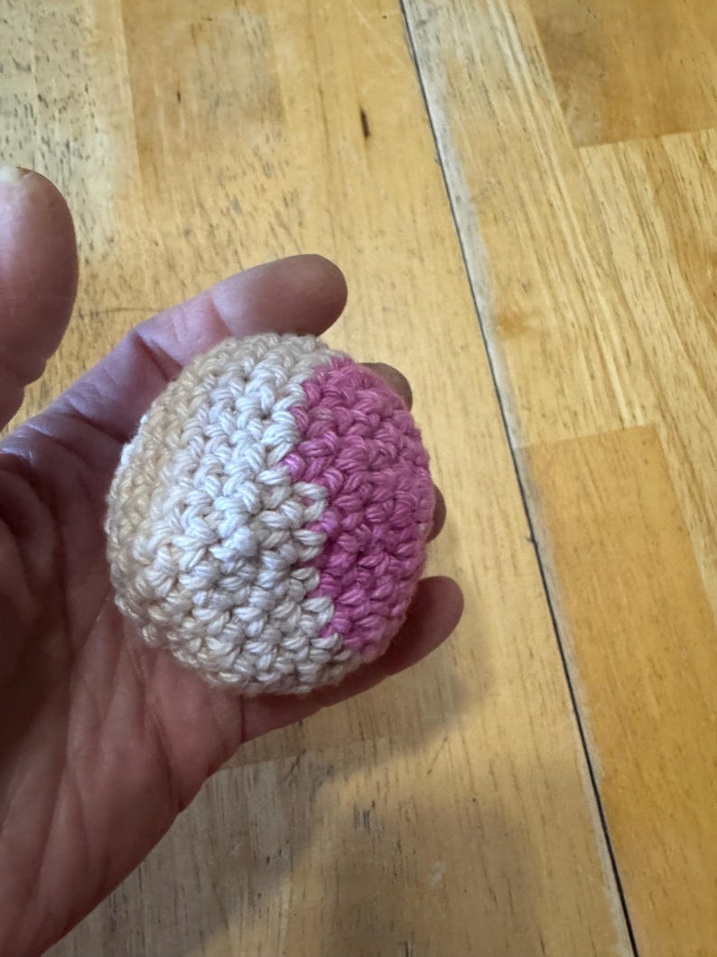 K&ouml;nnte beinhalten: Ein kleiner, handgefertigter H&auml;kelball in Beige-, Wei&szlig;- und Rosat&ouml;nen. Der Ball wird in einer Hand gehalten und scheint aus Garn zu bestehen. Die H&auml;kelstiche sind sichtbar, und der Ball hat eine runde Form.