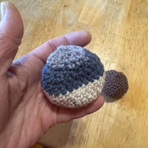 K&ouml;nnte beinhalten: Ein geh&auml;kelter Ball mit einem farbblockierten Design in Blau-, Grau- und Cremet&ouml;nen. Ein kleinerer, einfarbiger brauner geh&auml;kelter Ball ist ebenfalls sichtbar. Die Artikel sind handgefertigt und scheinen weich zu sein.