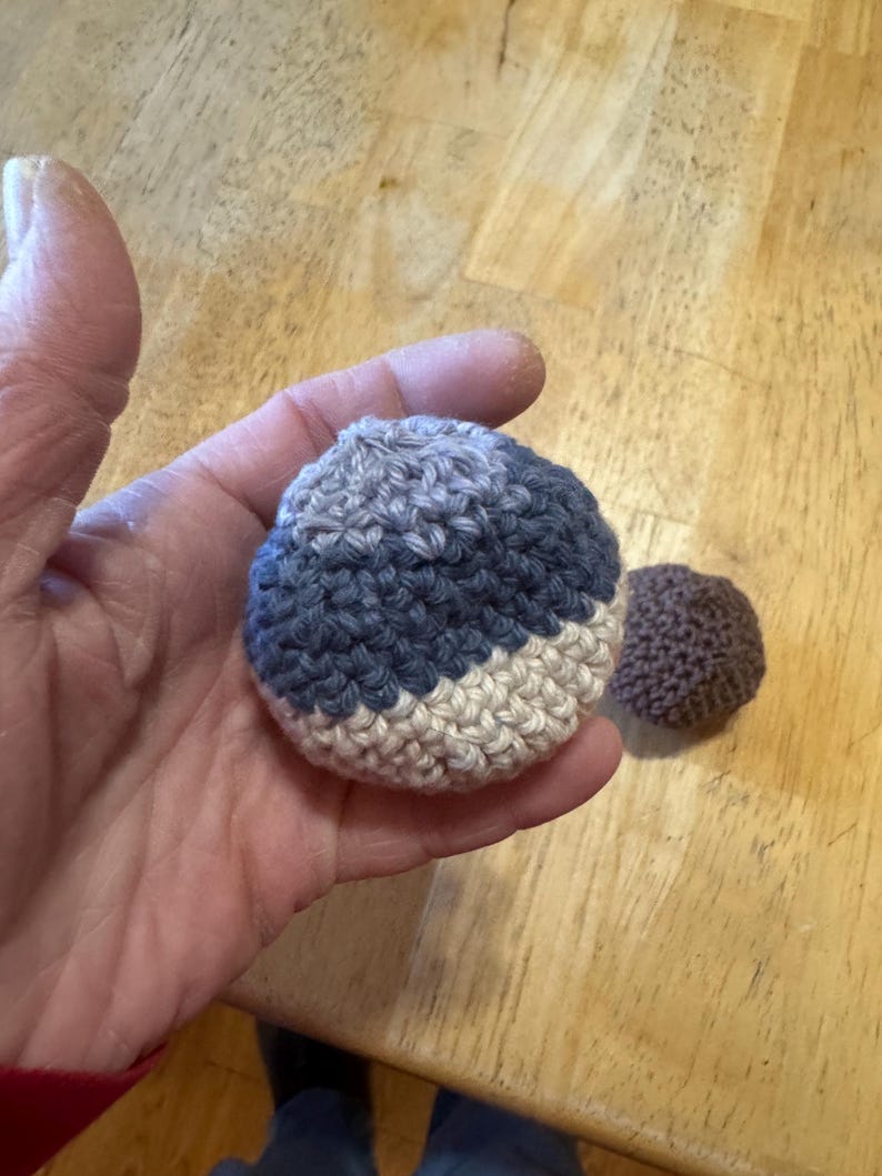 K&ouml;nnte beinhalten: Ein geh&auml;kelter Ball mit einem farbblockierten Design in Blau-, Grau- und Cremet&ouml;nen. Ein kleinerer, einfarbiger brauner geh&auml;kelter Ball ist ebenfalls sichtbar. Die Artikel sind handgefertigt und scheinen weich zu sein.