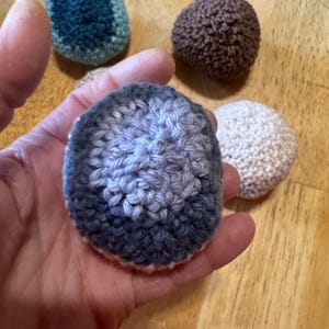 K&ouml;nnte beinhalten: Eine Sammlung handgefertigter, geh&auml;kelter Kugeln in verschiedenen Farben, darunter Blau, Braun und Creme. Eine Kugel wird in einer Hand gehalten, die ihre strukturierte Oberfl&auml;che und Farbvariationen zeigt. Die Kugeln sind auf einer Holzoberfl&auml;che angeordnet.