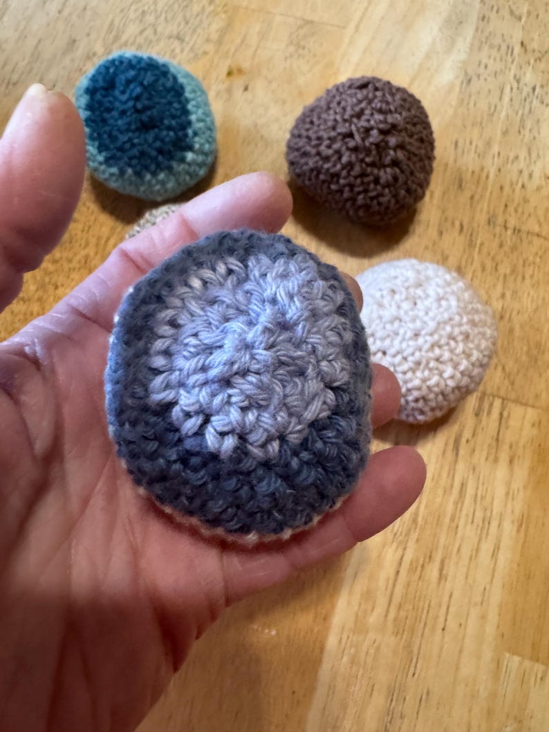K&ouml;nnte beinhalten: Eine Sammlung handgefertigter, geh&auml;kelter Kugeln in verschiedenen Farben, darunter Blau, Braun und Creme. Eine Kugel wird in einer Hand gehalten, die ihre strukturierte Oberfl&auml;che und Farbvariationen zeigt. Die Kugeln sind auf einer Holzoberfl&auml;che angeordnet.