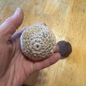 K&ouml;nnte beinhalten: Zwei geh&auml;kelte B&auml;lle, einer beige und einer braun, werden gezeigt. Der gr&ouml;&szlig;ere beige Ball wird in einer Hand gehalten, was die detaillierte Sticharbeit hervorhebt. Der kleinere braune Ball liegt daneben. Die B&auml;lle befinden sich auf einer Holzoberfl&auml;che.