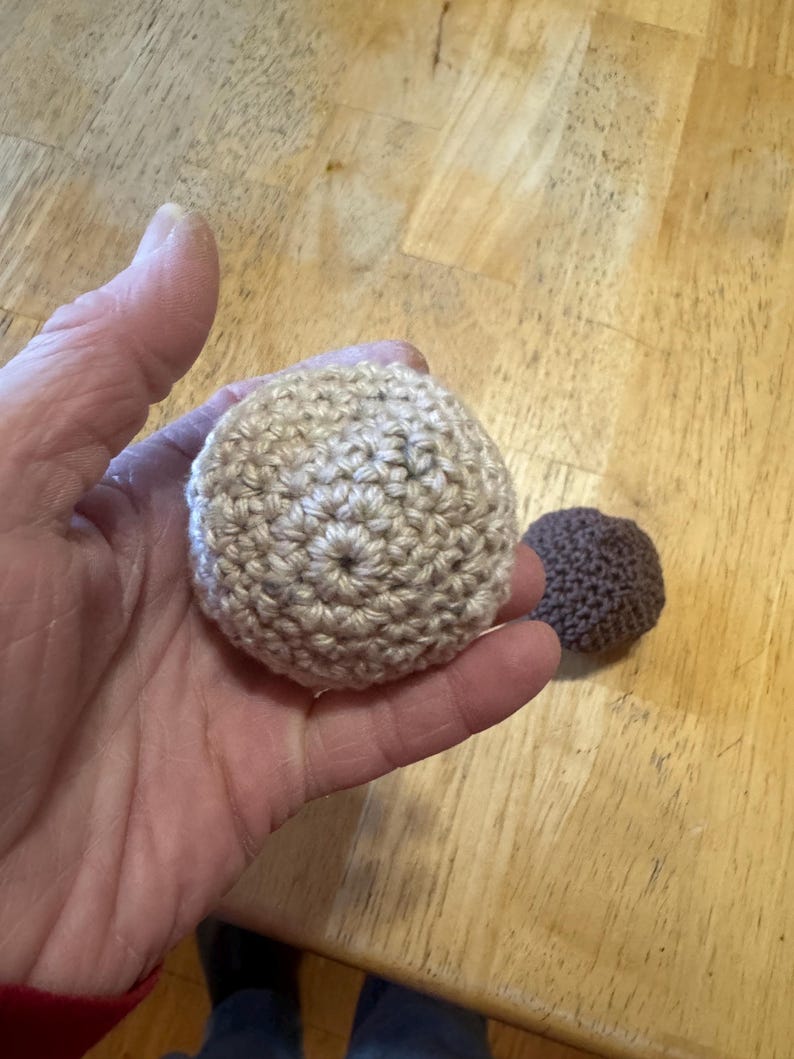K&ouml;nnte beinhalten: Zwei geh&auml;kelte B&auml;lle, einer beige und einer braun, werden gezeigt. Der gr&ouml;&szlig;ere beige Ball wird in einer Hand gehalten, was die detaillierte Sticharbeit hervorhebt. Der kleinere braune Ball liegt daneben. Die B&auml;lle befinden sich auf einer Holzoberfl&auml;che.