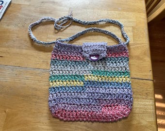 Könnte beinhalten: Handgehäkelte Umhängetasche in einer Vielzahl von Farben, darunter Rosa, Lila, Gelb und Grün. Die Tasche hat einen Klappenverschluss mit Knopf und einen geflochtenen Riemen. Die Tasche ist etwa 20 cm breit und 23 cm hoch.