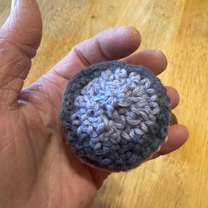 K&ouml;nnte beinhalten: Ein kleiner, runder, geh&auml;kelter Ball in Grau- und Blaut&ouml;nen. Der Ball wird in einer Hand gehalten und scheint handgefertigt zu sein. Die H&auml;kelstiche sind sichtbar und erzeugen eine strukturierte Oberfl&auml;che. Das Design des Balls hat eine hellere blaue Mitte.