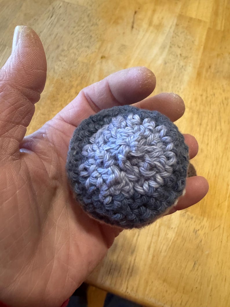 K&ouml;nnte beinhalten: Ein kleiner, runder, geh&auml;kelter Ball in Grau- und Blaut&ouml;nen. Der Ball wird in einer Hand gehalten und scheint handgefertigt zu sein. Die H&auml;kelstiche sind sichtbar und erzeugen eine strukturierte Oberfl&auml;che. Das Design des Balls hat eine hellere blaue Mitte.