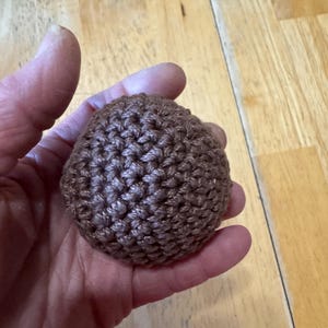 K&ouml;nnte beinhalten: Ein kleiner, brauner, geh&auml;kelter Ball. Der Ball wird in einer Hand gehalten und scheint aus einem strukturierten Garn hergestellt zu sein. Die Maschen sind sichtbar und erzeugen eine detaillierte Oberfl&auml;che. Die Farbe ist ein warmes, erdiges Braun.