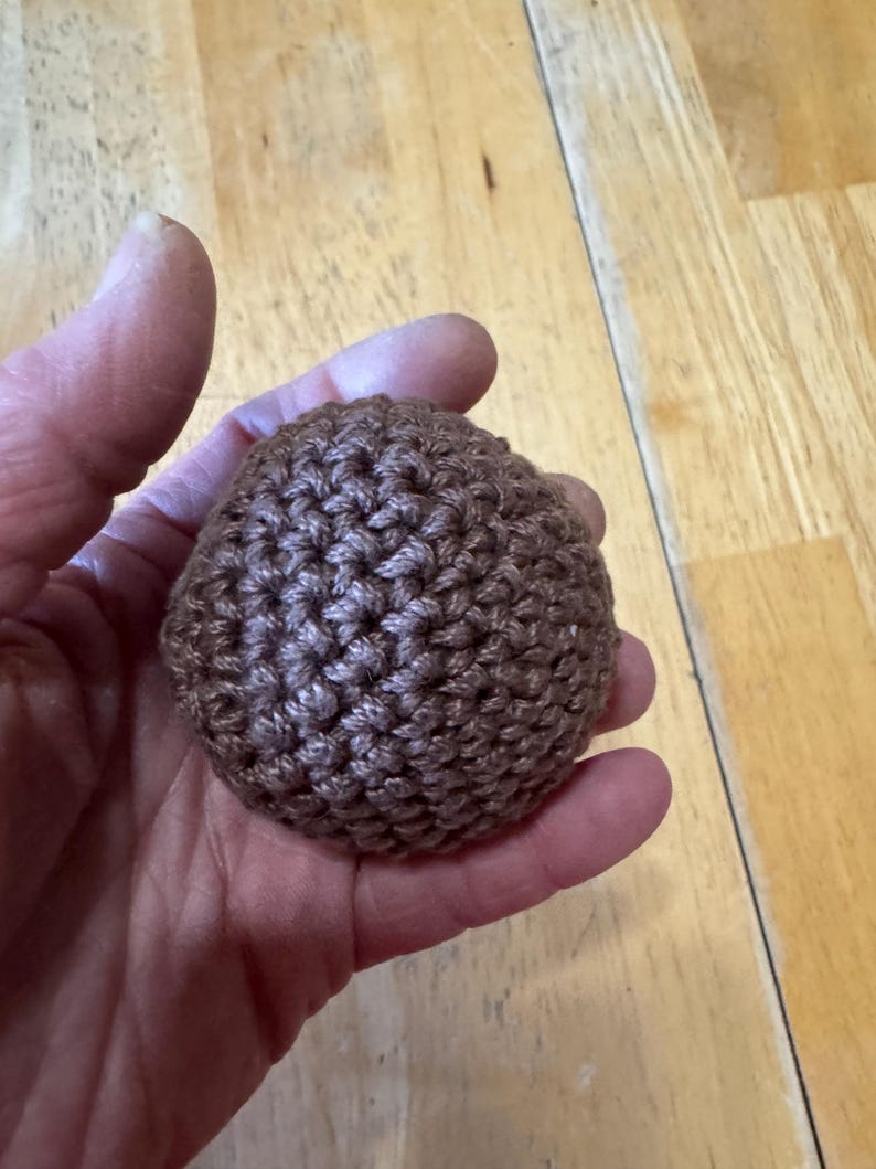 K&ouml;nnte beinhalten: Ein kleiner, brauner, geh&auml;kelter Ball. Der Ball wird in einer Hand gehalten und scheint aus einem strukturierten Garn hergestellt zu sein. Die Maschen sind sichtbar und erzeugen eine detaillierte Oberfl&auml;che. Die Farbe ist ein warmes, erdiges Braun.
