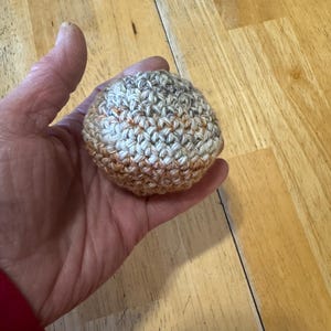 K&ouml;nnte beinhalten: Ein kleiner, runder, geh&auml;kelter Ball in Beige-, Braun- und Oranget&ouml;nen. Der Ball wird in einer Hand gehalten, wodurch die Textur und die Farbvariationen des Garns zur Geltung kommen. Der handgefertigte Artikel scheint ein Stressball oder ein dekorativer Gegenstand zu sein.