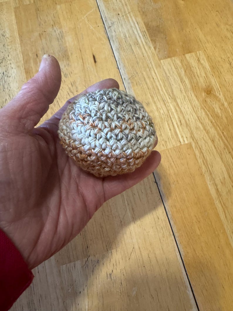K&ouml;nnte beinhalten: Ein kleiner, runder, geh&auml;kelter Ball in Beige-, Braun- und Oranget&ouml;nen. Der Ball wird in einer Hand gehalten, wodurch die Textur und die Farbvariationen des Garns zur Geltung kommen. Der handgefertigte Artikel scheint ein Stressball oder ein dekorativer Gegenstand zu sein.
