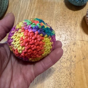 K&ouml;nnte beinhalten: Eine Hand h&auml;lt einen geh&auml;kelten Ball mit einer lebendigen Farbmischung. Der Ball besteht aus roter, orangefarbener, gelber, gr&uuml;ner, violetter und blauer Wolle. Weitere geh&auml;kelte B&auml;lle in Blau- und Gr&uuml;nt&ouml;nen sind im Hintergrund zu sehen.