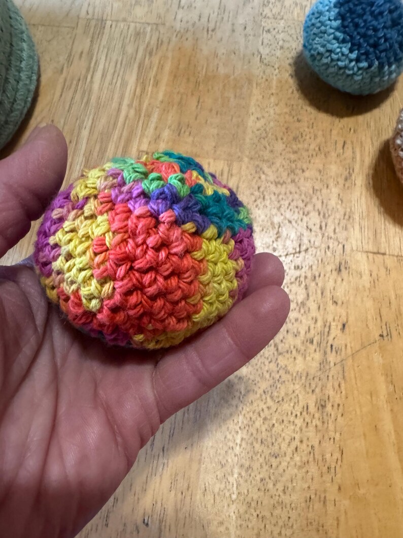 K&ouml;nnte beinhalten: Eine Hand h&auml;lt einen geh&auml;kelten Ball mit einer lebendigen Farbmischung. Der Ball besteht aus roter, orangefarbener, gelber, gr&uuml;ner, violetter und blauer Wolle. Weitere geh&auml;kelte B&auml;lle in Blau- und Gr&uuml;nt&ouml;nen sind im Hintergrund zu sehen.
