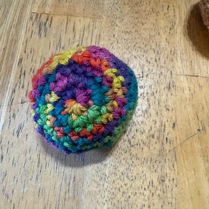K&ouml;nnte beinhalten: Ein handgeh&auml;kelter Ball in einer lebendigen Farbpalette aus Rot, Orange, Gelb, Gr&uuml;n, Blau und Lila. Der Ball ist rund und scheint aus weichem Garn gefertigt zu sein. Die Maschen sind sichtbar, was auf einen handgefertigten Artikel hindeutet.