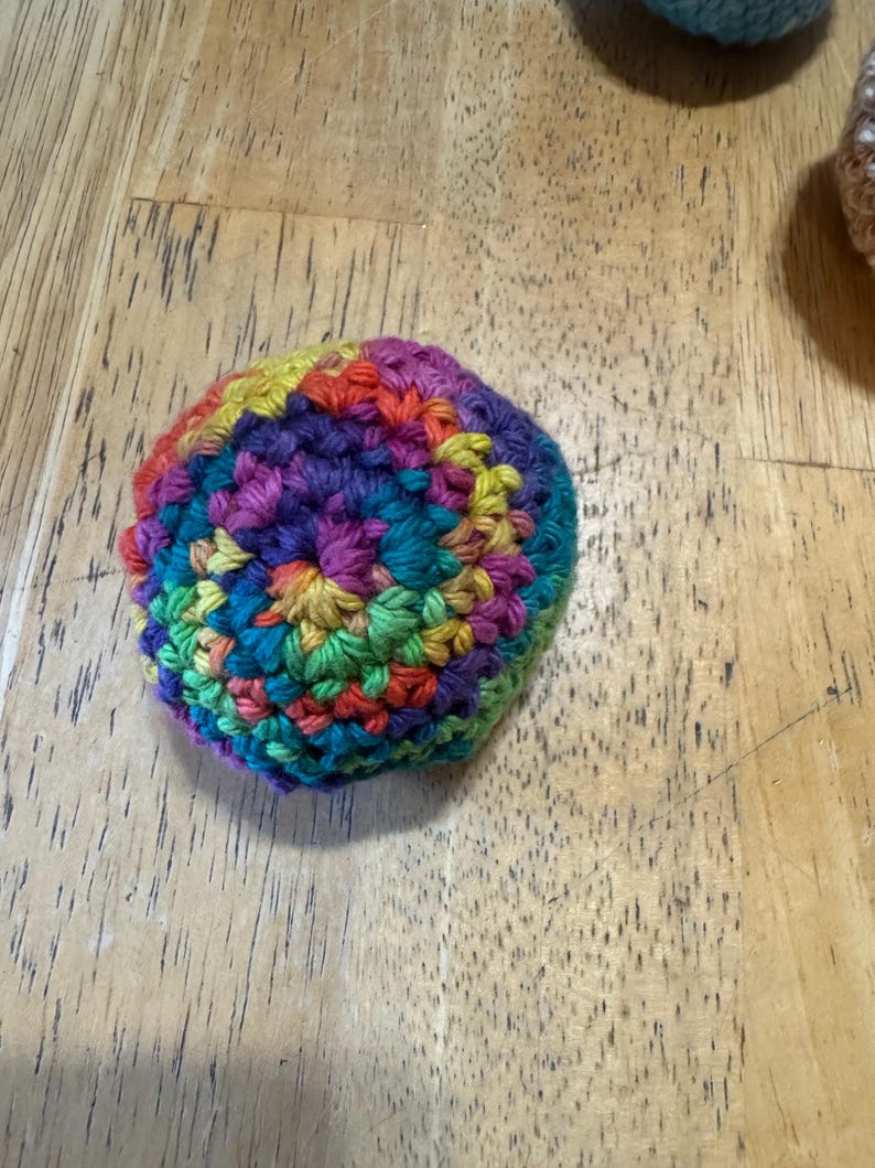 K&ouml;nnte beinhalten: Ein handgeh&auml;kelter Ball in einer lebendigen Farbpalette aus Rot, Orange, Gelb, Gr&uuml;n, Blau und Lila. Der Ball ist rund und scheint aus weichem Garn gefertigt zu sein. Die Maschen sind sichtbar, was auf einen handgefertigten Artikel hindeutet.