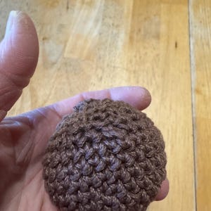 K&ouml;nnte beinhalten: Ein kleiner, brauner, geh&auml;kelter Ball, der in einer Hand gehalten wird. Der Ball hat eine strukturierte Oberfl&auml;che, die durch die engen Maschen entsteht. Die Farbe ist ein warmes, erdiges Braun, und die Gesamtform ist rund.