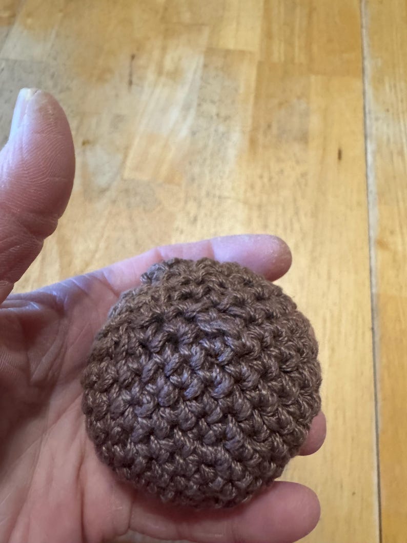 K&ouml;nnte beinhalten: Ein kleiner, brauner, geh&auml;kelter Ball, der in einer Hand gehalten wird. Der Ball hat eine strukturierte Oberfl&auml;che, die durch die engen Maschen entsteht. Die Farbe ist ein warmes, erdiges Braun, und die Gesamtform ist rund.