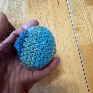K&ouml;nnte beinhalten: Ein kleiner, runder, geh&auml;kelter Ball in Blau- und T&uuml;rkist&ouml;nen. Das handgefertigte Objekt wird in einer Hand gehalten, wodurch die Textur des Garns und die engen Maschen hervorgehoben werden. Der Ball scheint ein Bastelartikel oder ein Spielzeug zu sein.