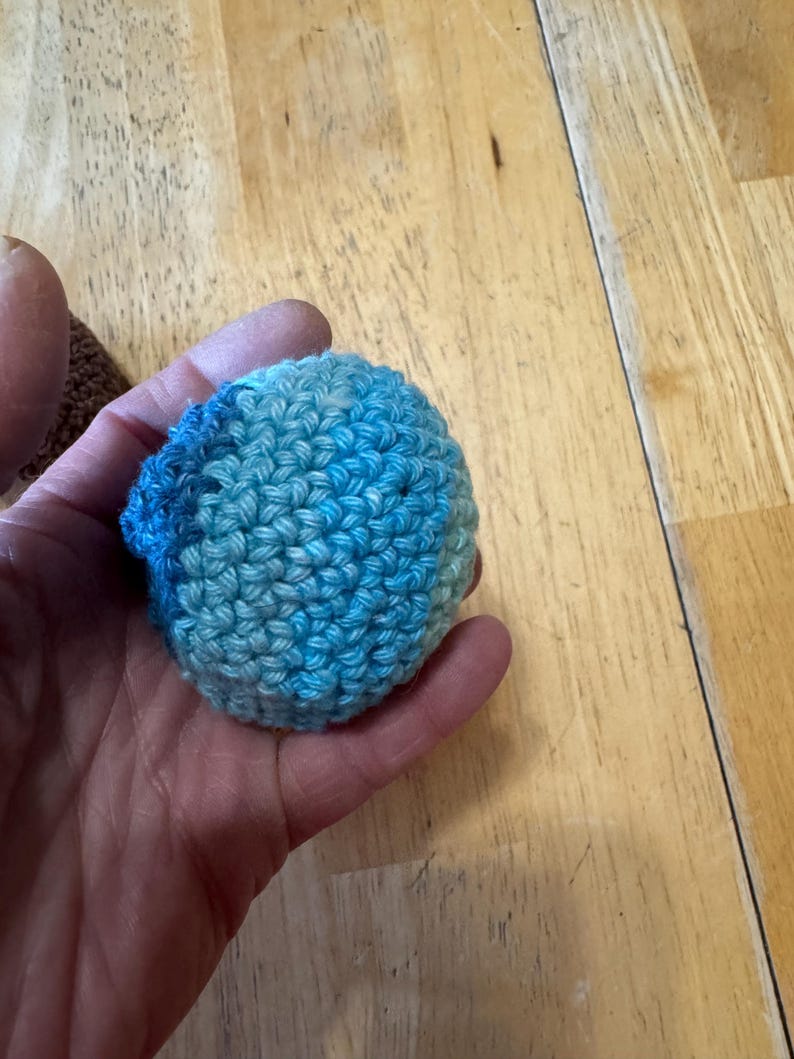 K&ouml;nnte beinhalten: Ein kleiner, runder, geh&auml;kelter Ball in Blau- und T&uuml;rkist&ouml;nen. Das handgefertigte Objekt wird in einer Hand gehalten, wodurch die Textur des Garns und die engen Maschen hervorgehoben werden. Der Ball scheint ein Bastelartikel oder ein Spielzeug zu sein.