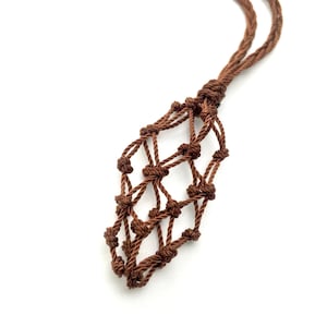Op de afbeelding: Een bruine macram&eacute; ketting met een geweven zakje dat is ontworpen om een klein voorwerp in te bewaren. Het koord van de ketting is gedraaid en geknoopt, en het zakje is een ruitvormig net. De achtergrond is wit.