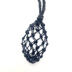 May include: A black macrame crystal holder with a woven net design. The holder has a long, braided cord for wearing around the neck. The macrame is knotted to create a pocket to hold a crystal or other small object.