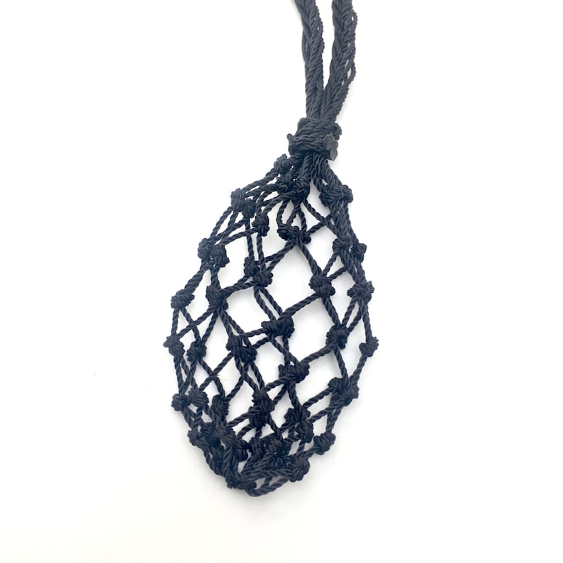 May include: A black macrame crystal holder with a woven net design. The holder has a long, braided cord for wearing around the neck. The holder is designed to hold a crystal or other small object.