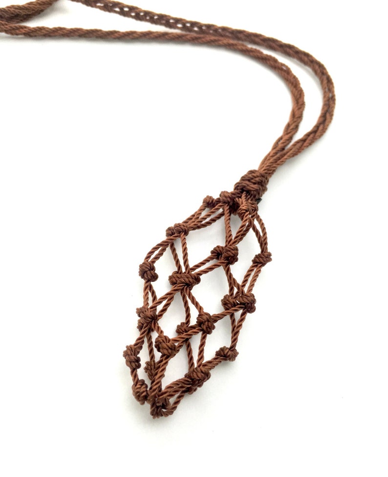 Op de afbeelding: Een bruine macram&eacute; ketting met een geweven zakje dat is ontworpen om een klein voorwerp in te bewaren. Het koord van de ketting is gedraaid en geknoopt, en het zakje is een ruitvormig net. De achtergrond is wit.