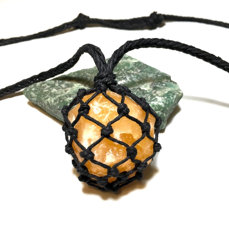 May include: A handmade necklace featuring a golden-orange crystal pendant encased in a black macrame net. The necklace cord is also black. The pendant rests on a green stone against a white background.