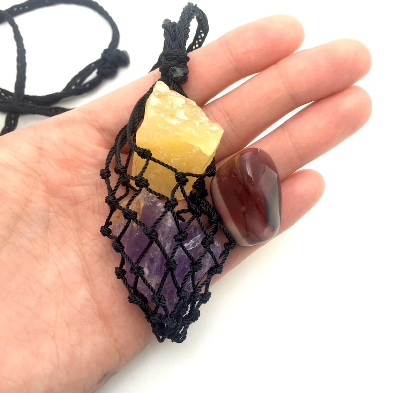 May include: A black macrame necklace featuring three raw gemstones. The necklace holds a yellow calcite crystal, a purple amethyst crystal, and a polished red jasper stone. The gemstones are displayed against a white background.