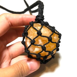 May include: A golden-yellow crystal pendant encased in a black woven net, suspended from a black cord. The crystal has a rough, natural texture. The cord is knotted at the top for wearing. The pendant is held in a hand.