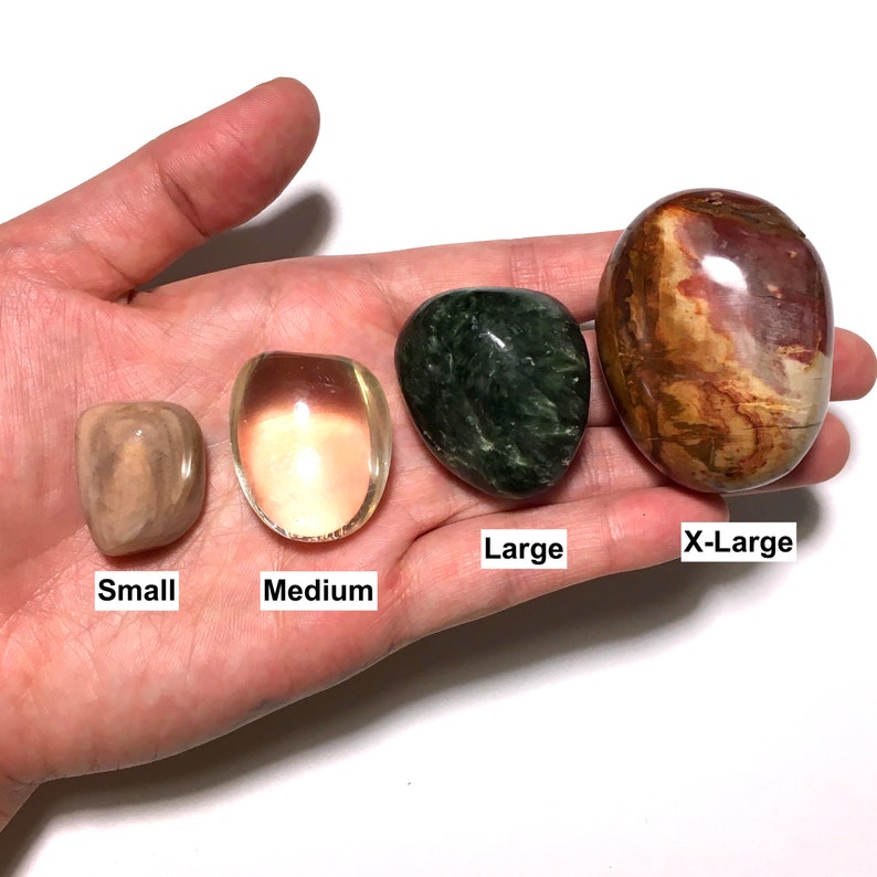 Op de afbeelding: Vier gepolijste stenen van verschillende formaten, gelabeld met "Small", "Medium", "Large" en "X-Large". De stenen vertonen een reeks kleuren, waaronder beige, helder met een perzikkleur, donkergroen en bruin met rode en bruine strepen. De stenen worden in een hand gehouden.