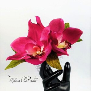 May include: A pink orchid hair comb with a pearl accent. The comb is held in a black hand.