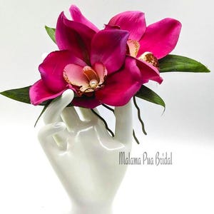 May include: Two pink orchid hair clips with pearl accents. The clips are held in a white hand.