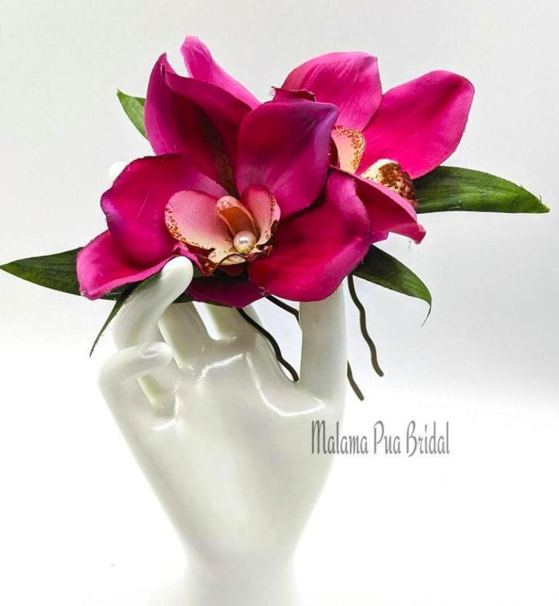 May include: Two pink orchid hair clips with pearl accents. The clips are held in a white hand.