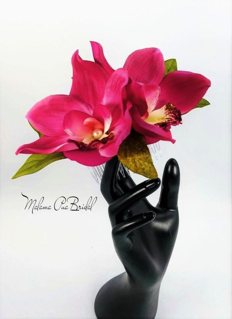 May include: A pink orchid hair comb with a pearl accent. The comb is held in a black hand.