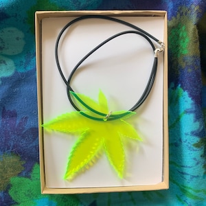 May include: A green and yellow leaf-shaped pendant necklace with a black cord. The necklace is displayed in a small cardboard box.
