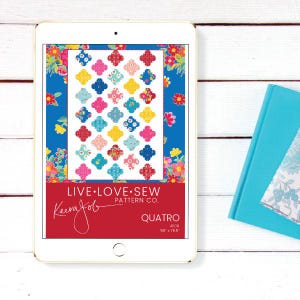May include: A digital tablet shows a quilt pattern with a repeating four-leaf clover design in pink, blue, yellow, and red. The tablet screen displays the text "LIVE.LOVE.SEW" and "QUATRO". The quilt measures 139.7cm x 194.3cm. A blue book is visible on the right.