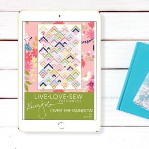 May include: A digital tablet displays a colorful quilt pattern titled "Over the Rainbow" by Keera Job. The pattern features geometric shapes in various colors. The tablet is next to a teal book on a white wooden surface.
