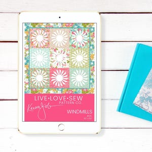 May include: A tablet shows a quilt pattern called "Windmills" by Keera Job. The quilt design has nine squares with a white sunburst on a floral background. The tablet is on a white wooden surface, with a teal book nearby. The quilt measures 127cm x 127cm.