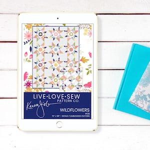 May include: A digital tablet displays a quilt pattern titled "Wildflowers" by Live Love Sew Pattern Co. The quilt design features star and floral motifs in pastel colors. The tablet is next to a teal book.