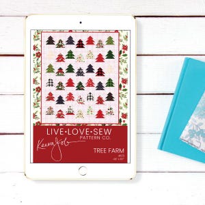 May include: A digital tablet displays a quilt pattern titled "Tree Farm" by Keera Job. The pattern features a grid of Christmas trees in various colors and patterns. The tablet is on a white wooden surface, with a blue book visible.