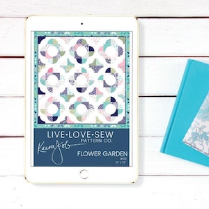 May include: A digital tablet displays a quilt pattern titled "Flower Garden" by Keera Job, with dimensions of 72" x 72". The quilt design features floral shapes in pastel colors. A teal book and fabric sample are visible on the right.