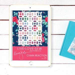Peut inclure: Une tablette numérique affiche un patron de courtepointe avec un motif floral et le texte "LIVE LOVE SEW". Le motif est un patchwork de carrés colorés. En dessous, une bannière rose avec le texte "CHAIN REACTION" et le nom du créateur.